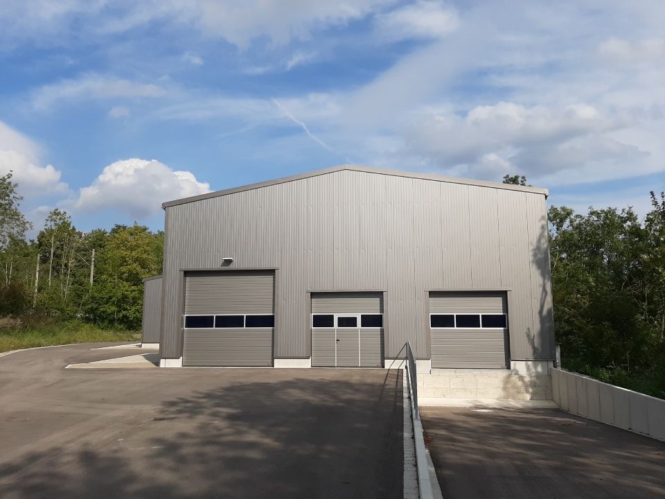 Graues Industriegebäude mit drei Garagentoren und einer gepflasterten Zufahrt unter einem teilweise bewölkten Himmel