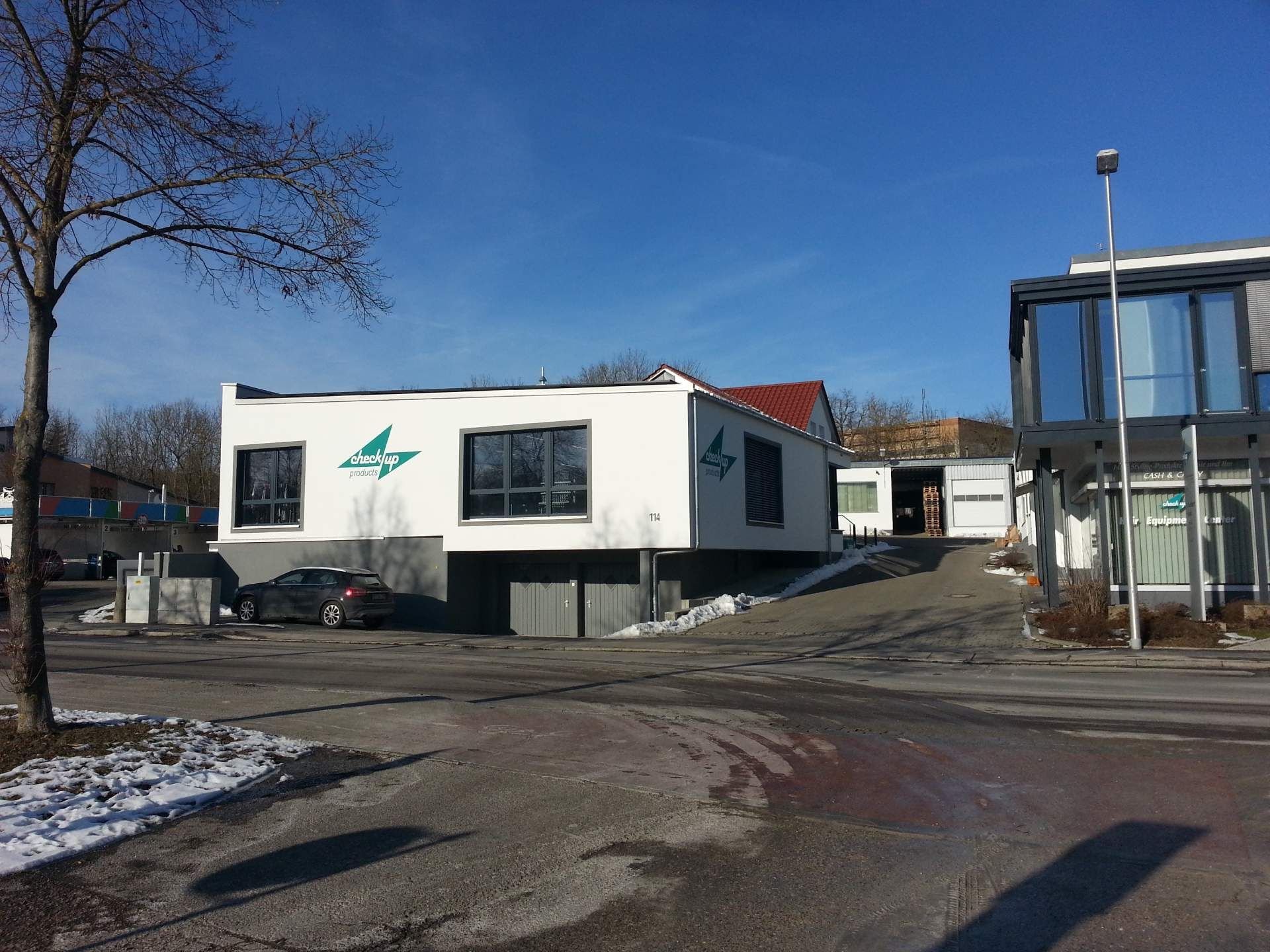 Kleines weißes Modulgebäude mit grünem Logo neben einem Glasbauwerk in einer verschneiten Straße.