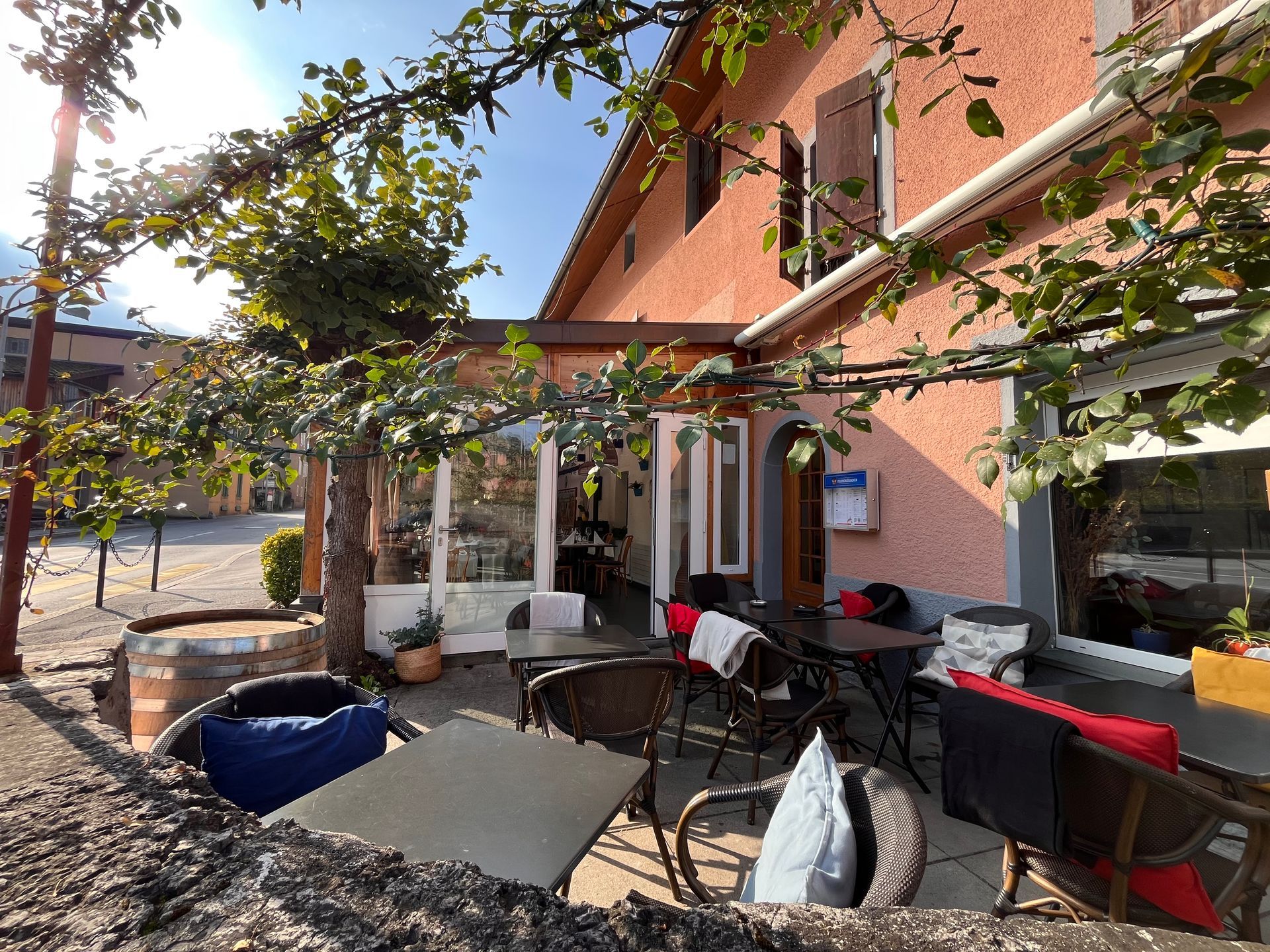 Terrasse extérieure avec tables et chaises devant un bâtiment rose, ombragée par des vignes.