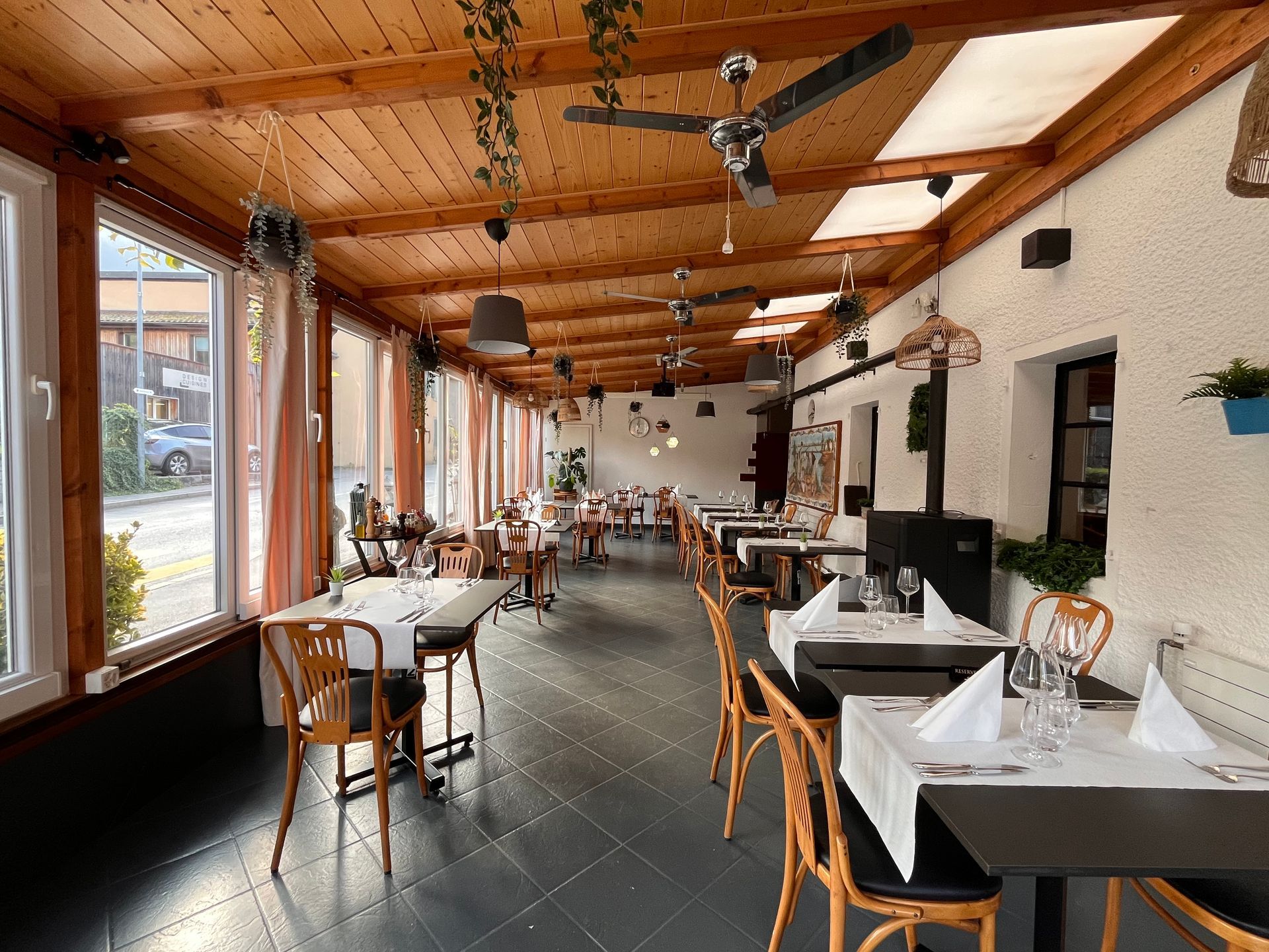 Intérieur de restaurant avec tables dressées pour dîner, plafond en bois et grandes fenêtres.