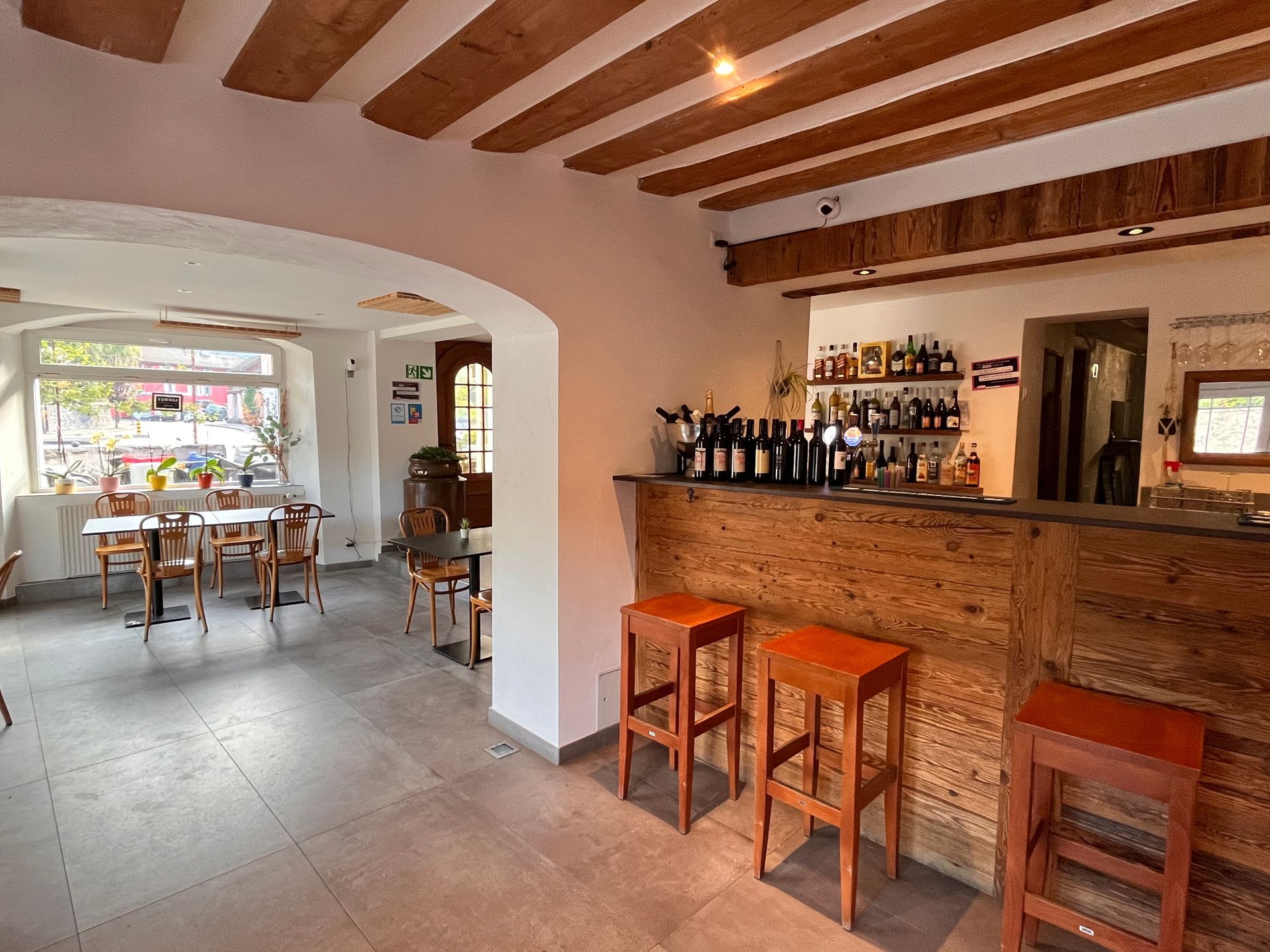 Vue intérieure d'un bar rustique avec poutres en bois, un bar, des tabourets et un petit coin repas avec des tables.