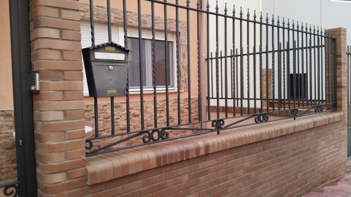 Una pared de ladrillos con una valla de hierro forjado y un buzón sobre ella.