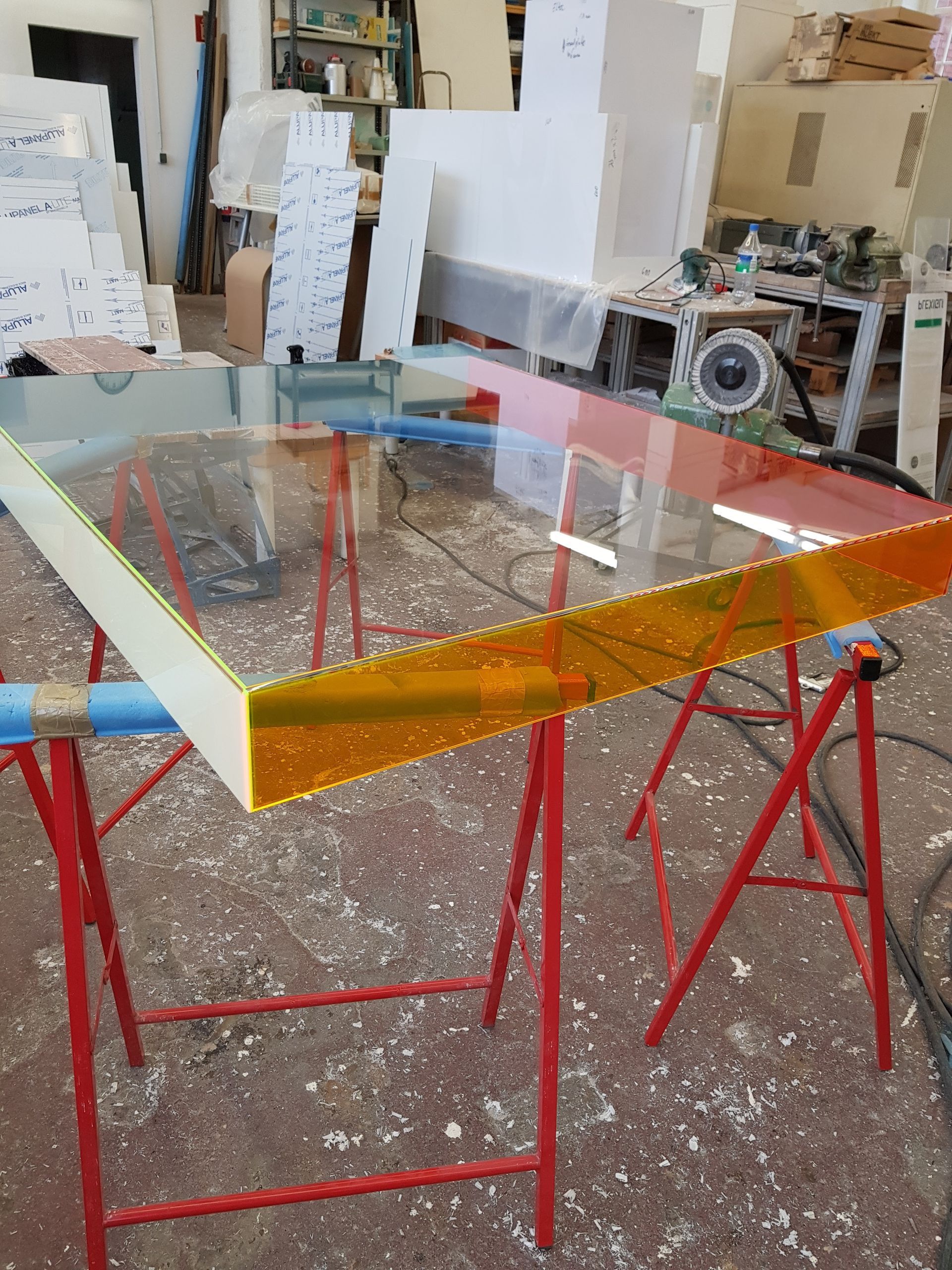 Große, transparente Glas- oder Acrylplatte auf Böcken in einer Werkstatt, mit Materialien im Hintergrund.