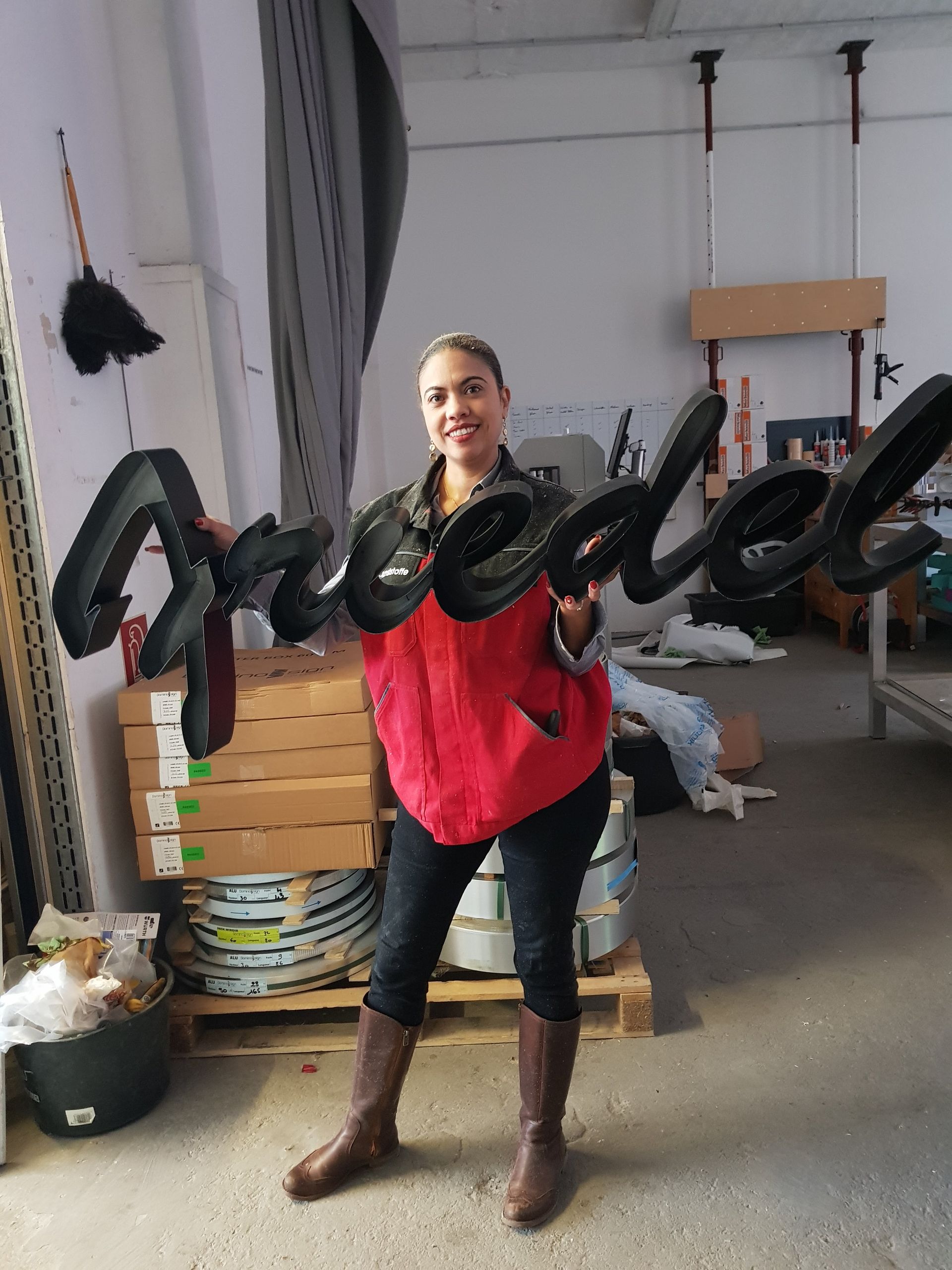 Mitarbeiterin steht neben großen, schwarzen Einzelbuchstaben in einer Werkstatt.