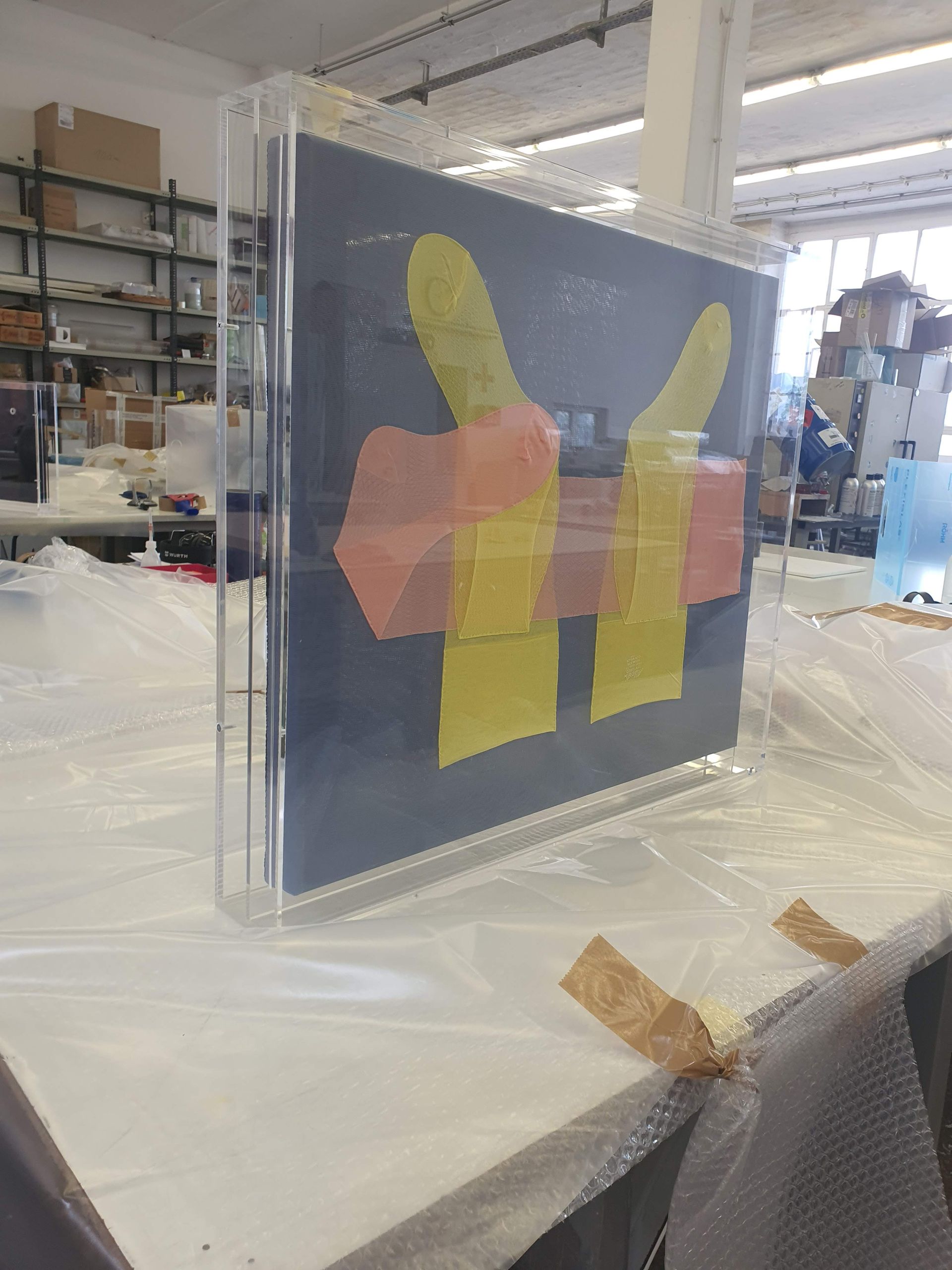 Transparente Platte mit farbigen, abstrakten Formen vor einem Werkstattregal.