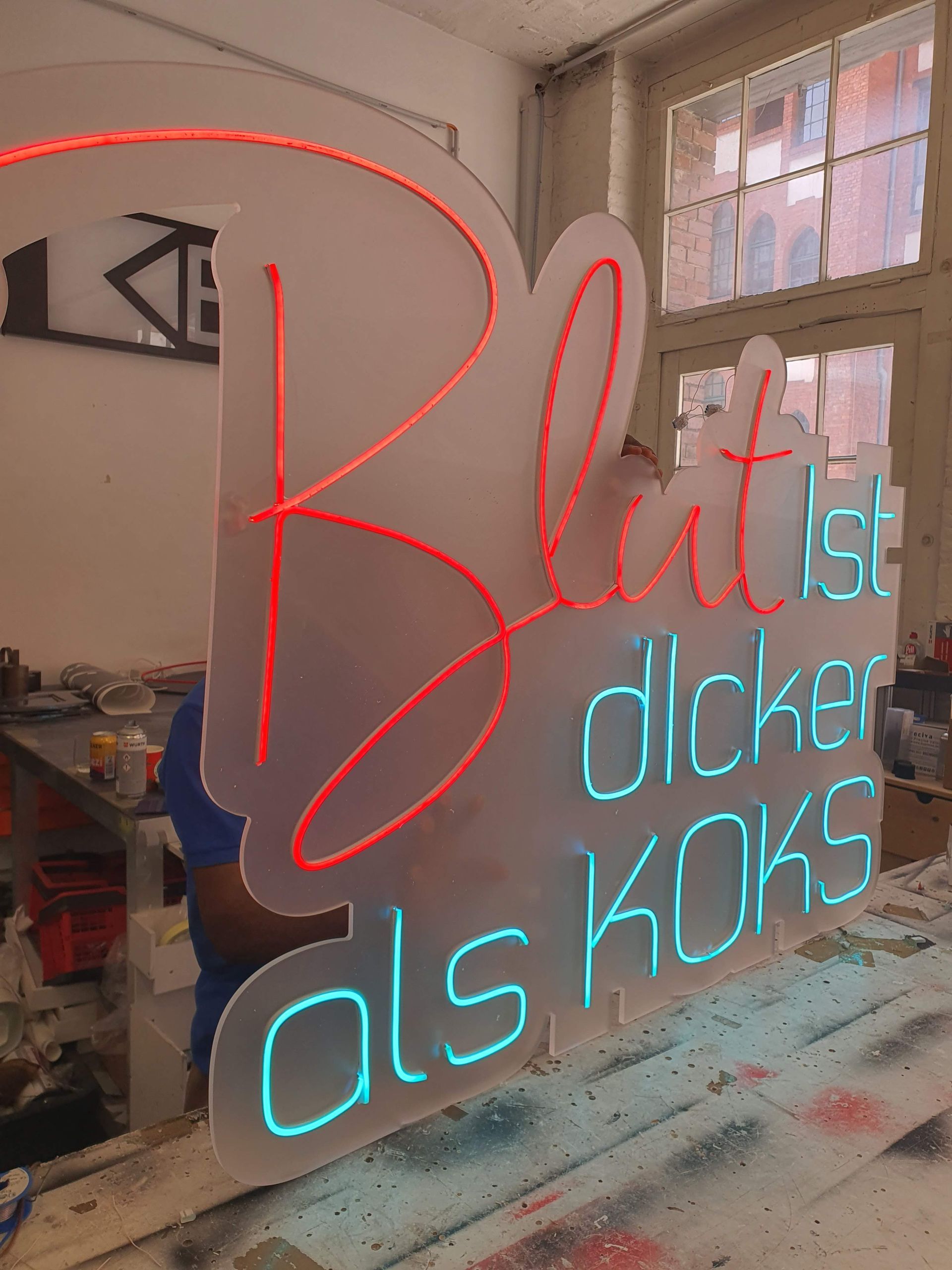 Roter Neon-Schriftzug an einer Wand in einer Werkstatt mit Fenstern im Hintergrund.
