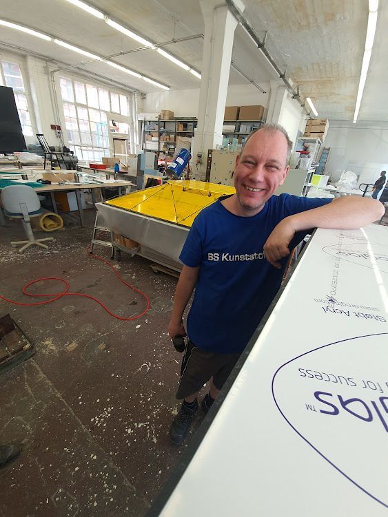 Lachender Mitarbeiter lehnt sich an eine gelbe Kunststoffkonstruktion in der Werkhalle.