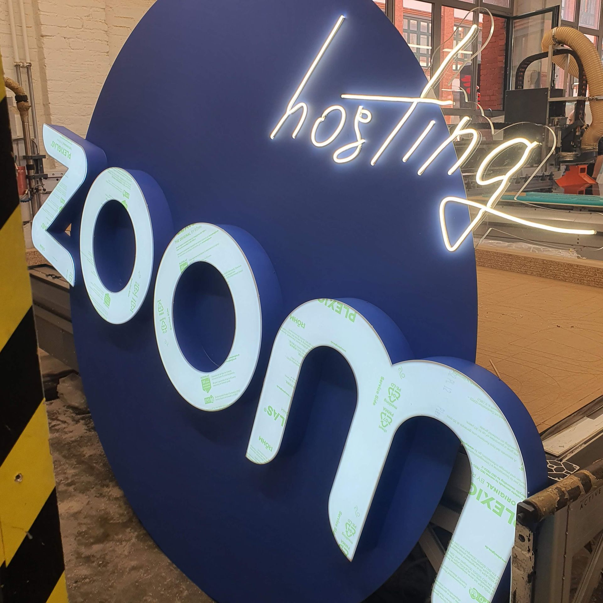 Neon-Schriftzug „Zoom hosting“ in Weiß und Blau, montiert in einer Werkstattumgebung.