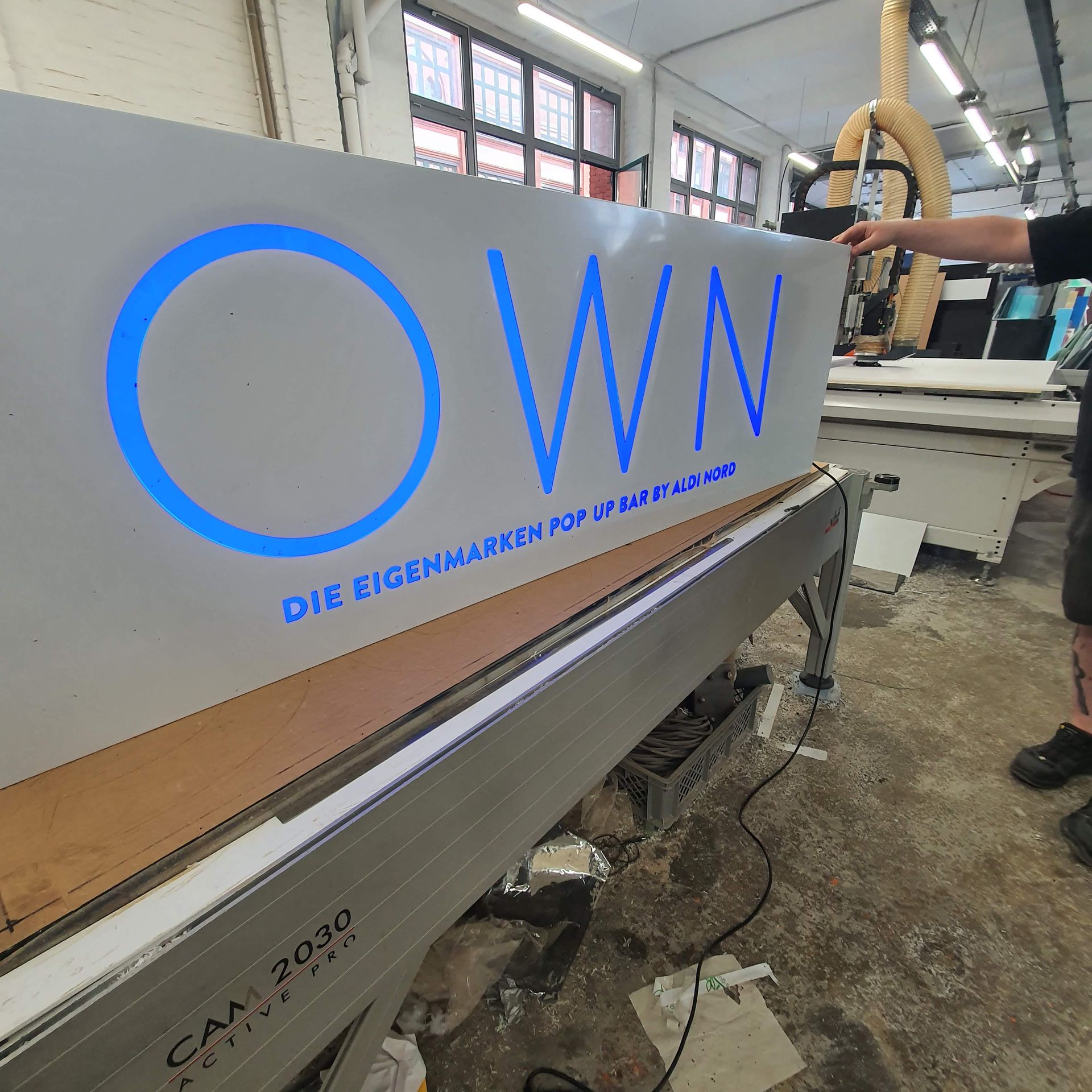 Graue Wandtafel mit blauem Schriftzug „OWN“ in einer Werkstatt.