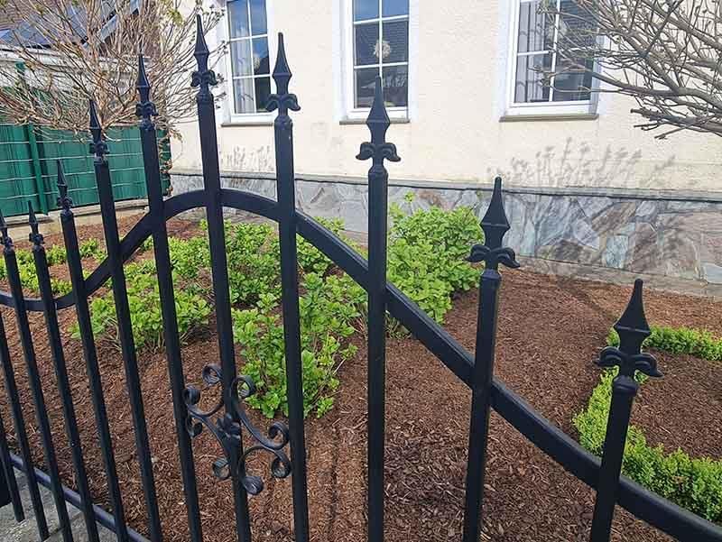 Ein schwarzer schmiedeeiserner Zaun umgibt einen Garten vor einem Haus.