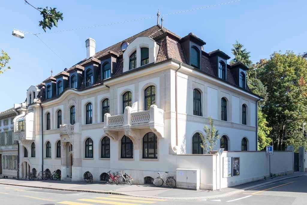 Weißes, mehrstöckiges Herrenhaus mit dunklem Dach und Balkonen an einer sonnigen Straßenecke