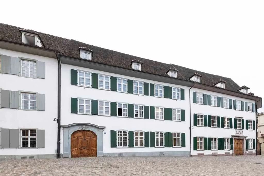 Eine Reihe weißer Gebäude mit grünen Fensterläden und einem gepflasterten Innenhof unter einem bewölkten Himmel