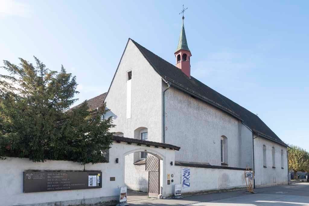 Weiße Kirchenfassade mit kleinem Kirchturm neben einer Straße und Bäumen unter blauem Himmel