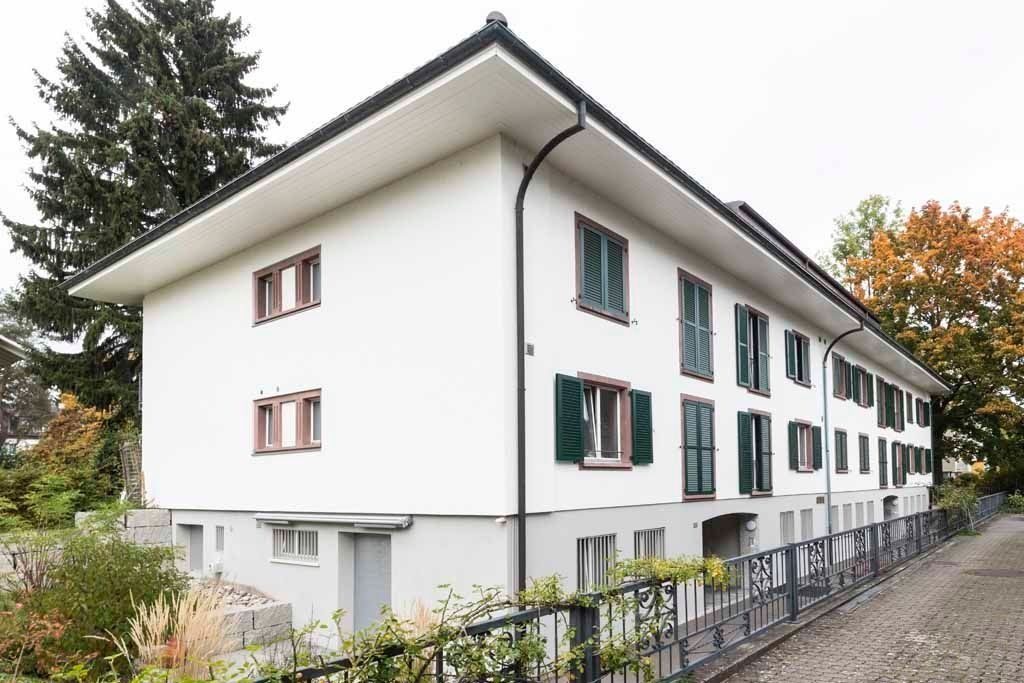 Weißes Mehrfamilienhaus mit grünen Fensterläden neben einem Fußweg und Bäumen
