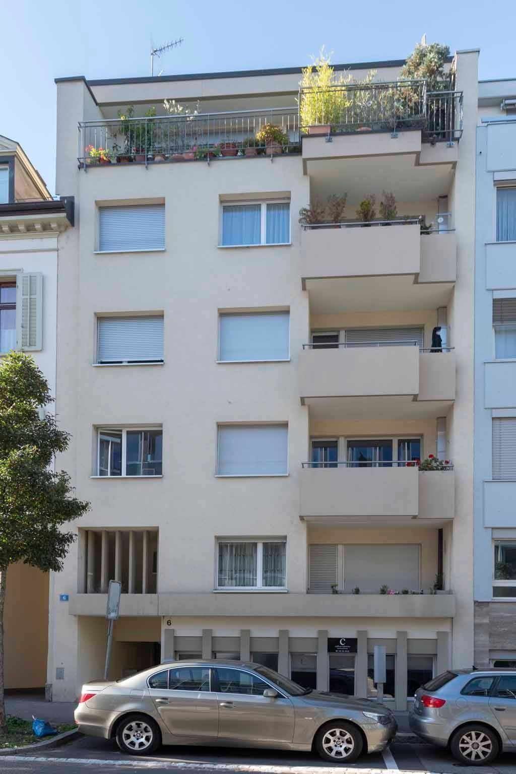 Fünfstöckiges Wohnhaus mit Balkonen und Topfpflanzen, davor parkende Autos.