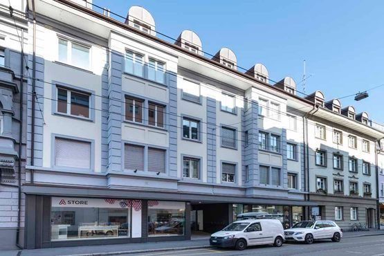 Straßenansicht eines hellgrauen Wohnhauses mit Ladenfronten und geparkten Autos unter klarem, blauem Himmel