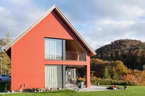 Rotes Haus mit steilem Dach und Balkon in einem grasbewachsenen Hof unter bewölktem Himmel.