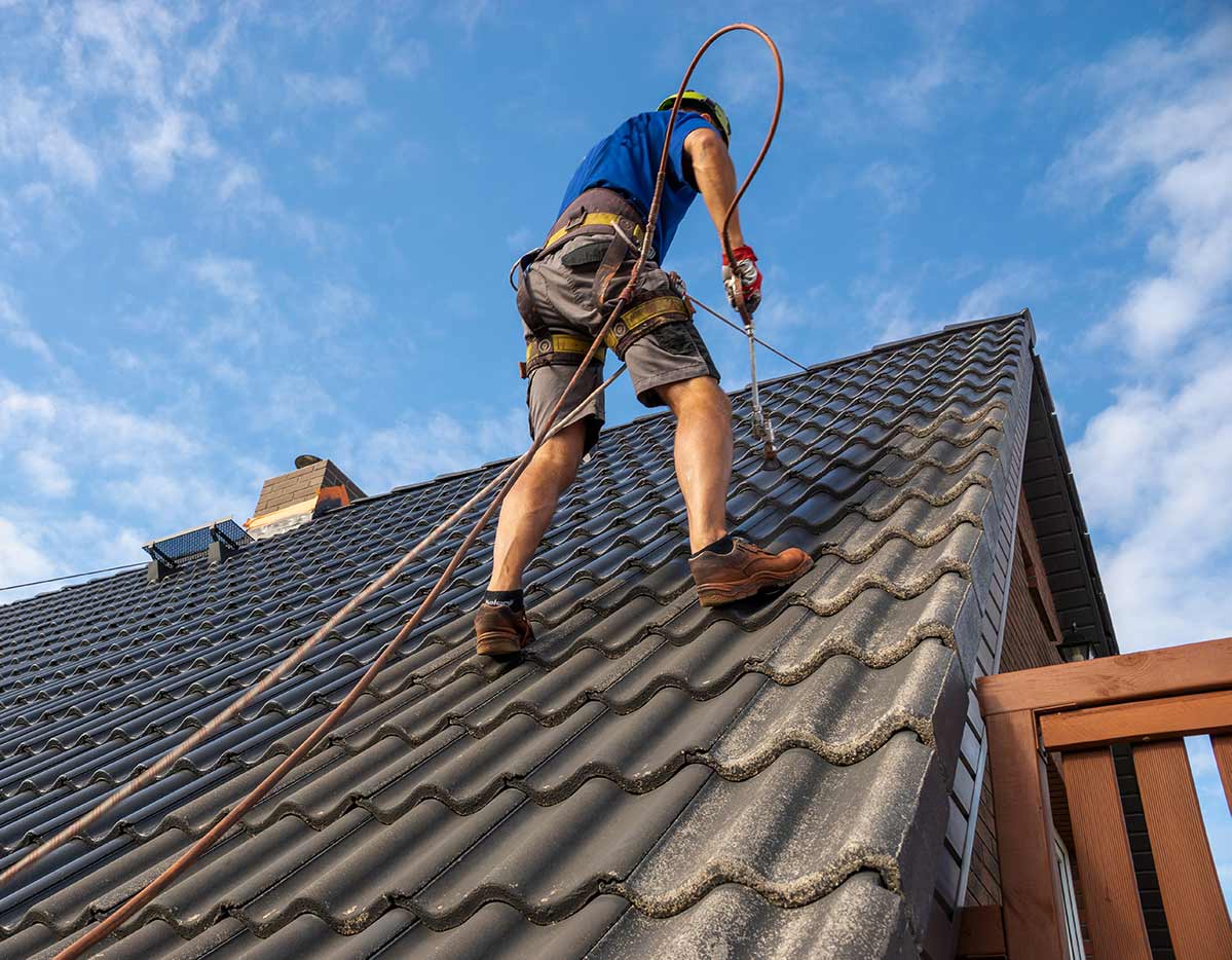 Un couvreur sur un toit de tuiles, sécurisé par un harnais et une corde, sous un ciel bleu.