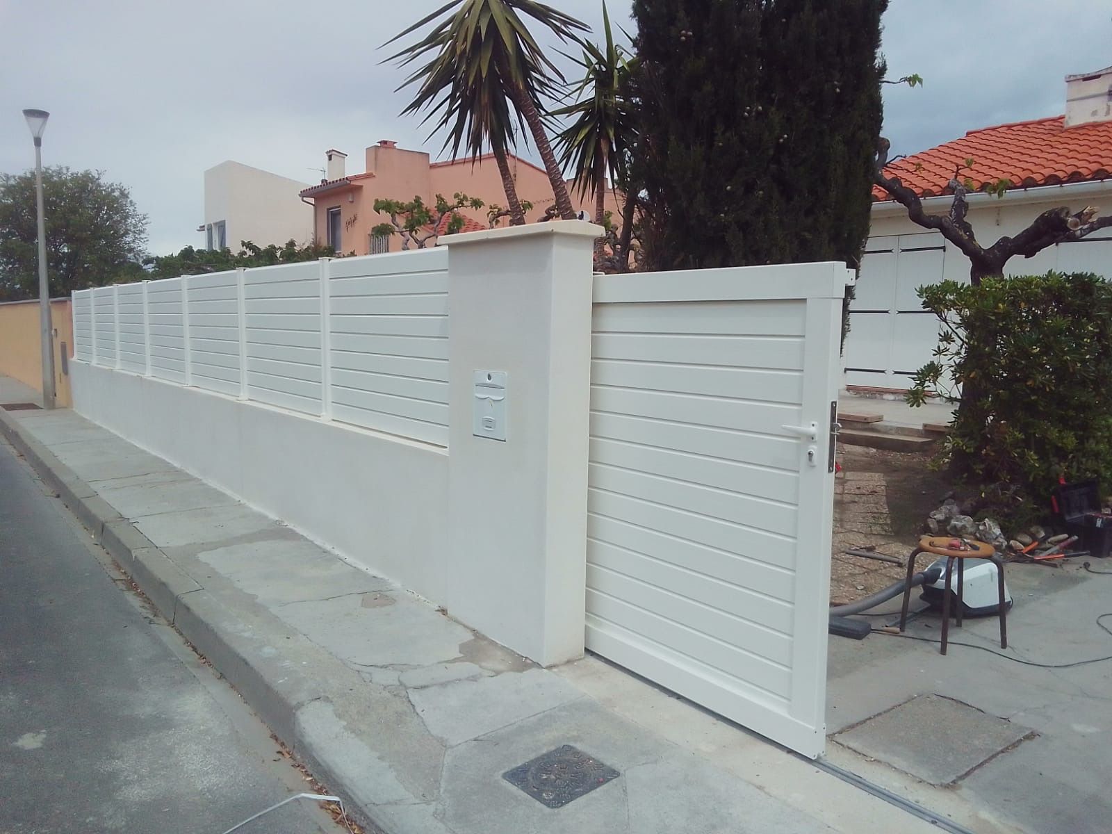 Clôture et portail blancs à claire-voie le long d'un trottoir, avec des bâtiments et des arbres en arrière-plan.