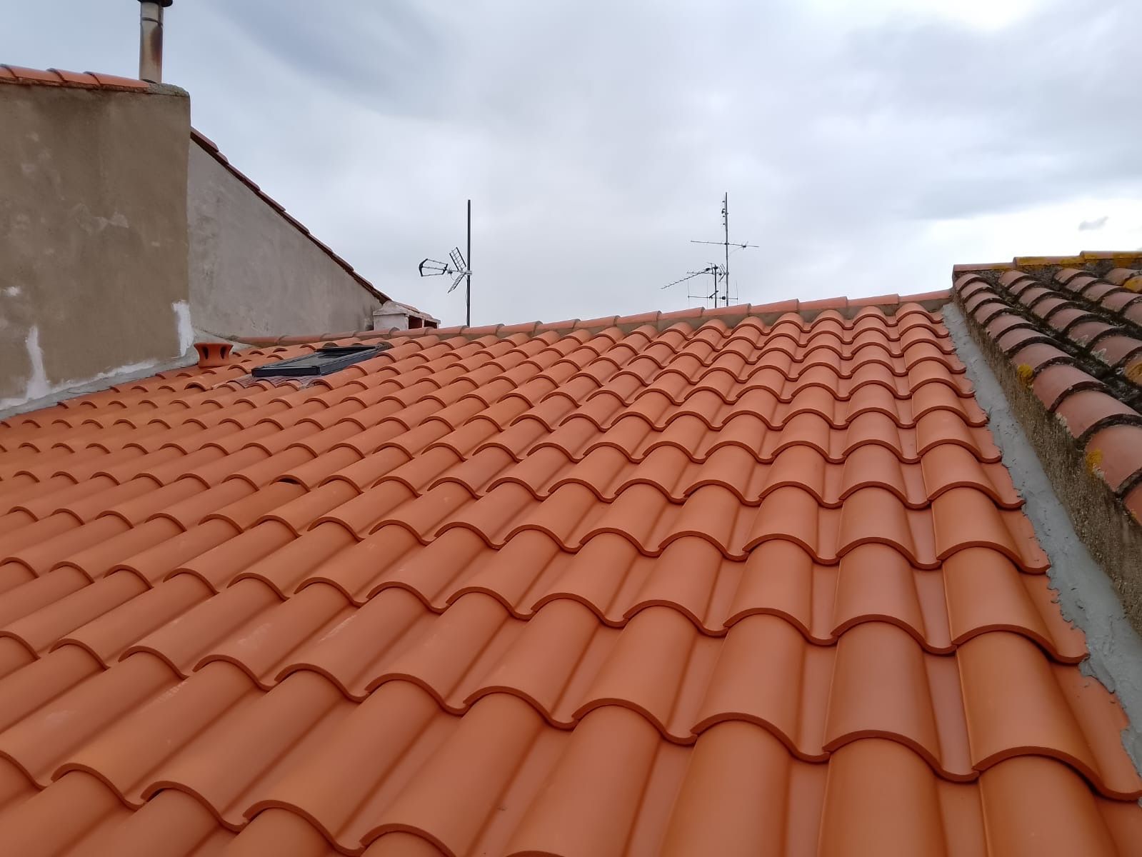 Toit de tuiles orange sous un ciel nuageux, avec une cheminée et des antennes visibles.