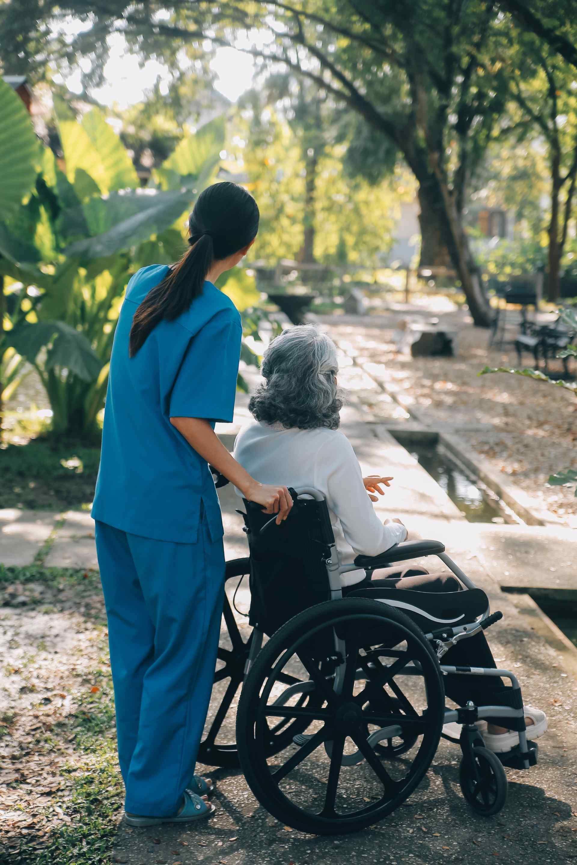 Eine Krankenschwester steht neben einer älteren Frau im Rollstuhl. Pflegedaheim24h
