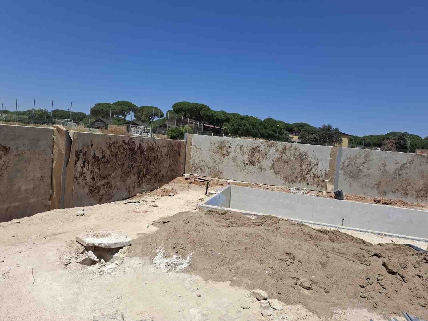 Obra en construcción con muros de cimentación de hormigón, montones de tierra y un cielo azul despejado.