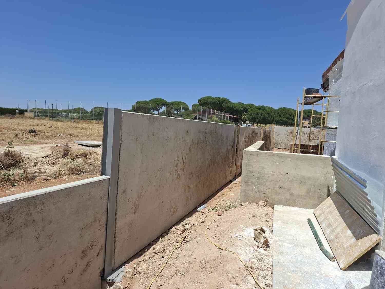 Un estrecho sendero de hormigón sin terminar junto a un muro, con un terreno baldío seco y el cielo azul al fondo.