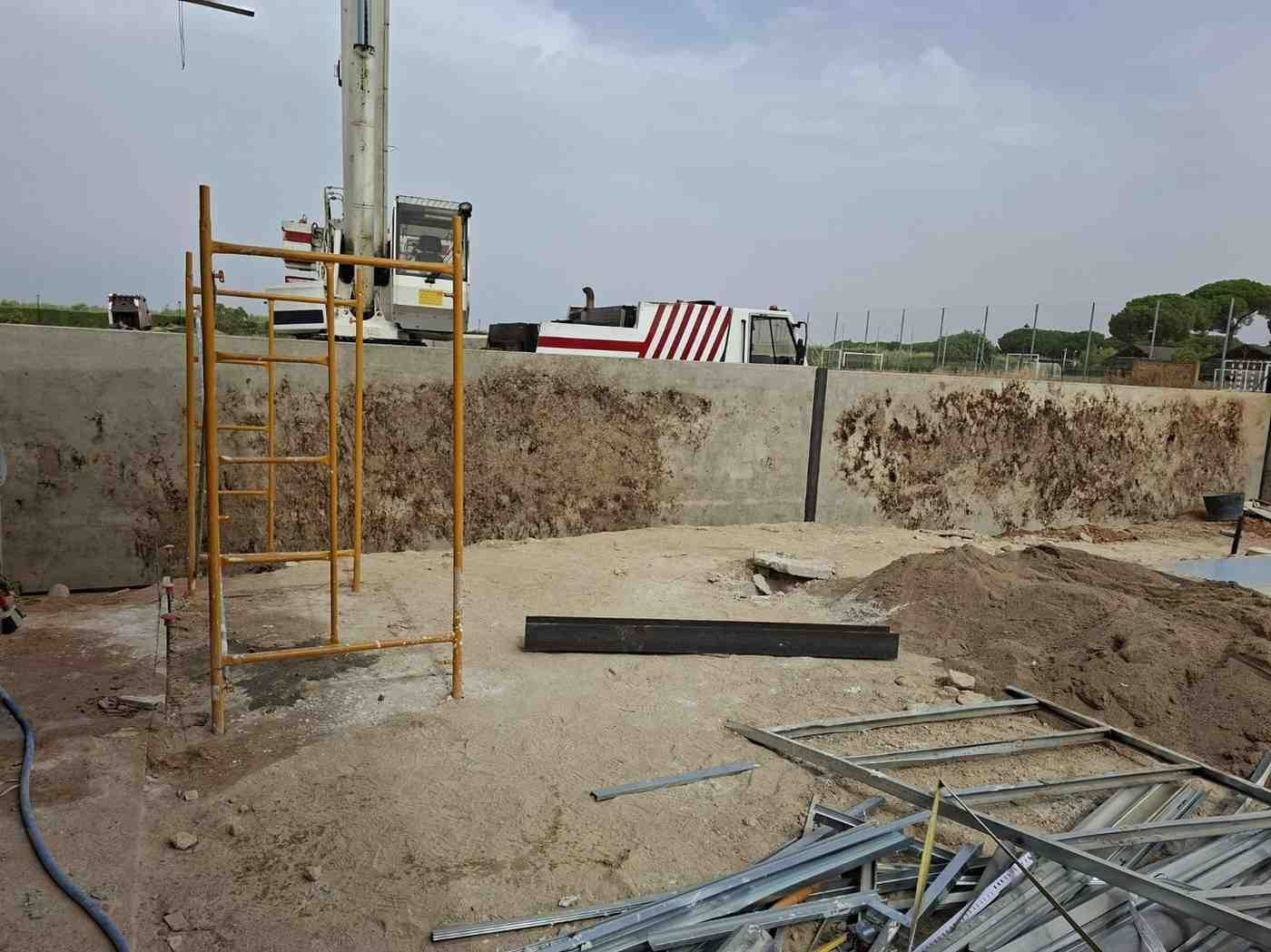 Obra en construcción con una excavación abierta, andamios metálicos y materiales apilados bajo un cielo nublado.