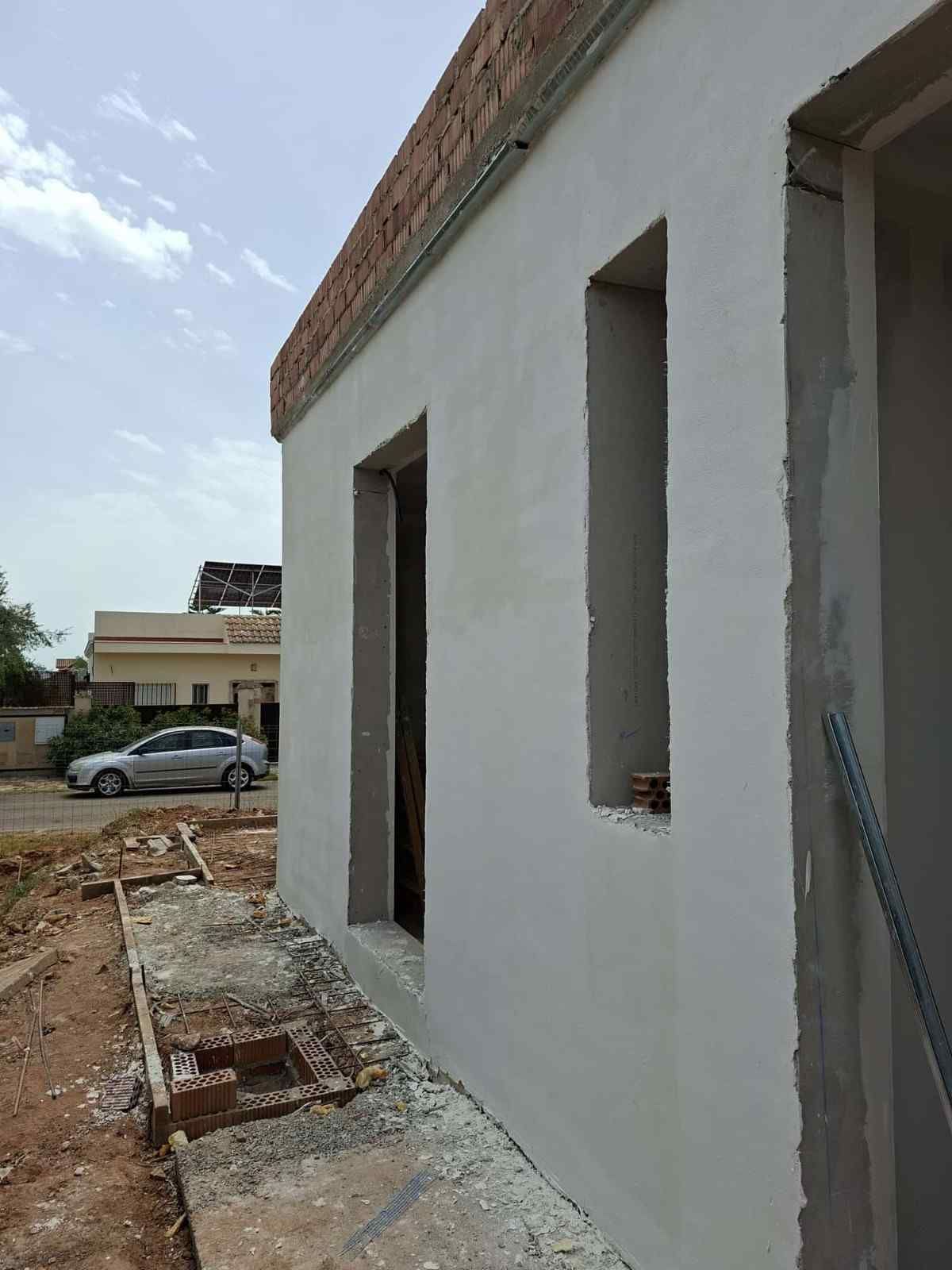 Muro blanco sin terminar de un edificio con estrechas aberturas para ventanas, junto a un terreno baldío y un coche aparcado.