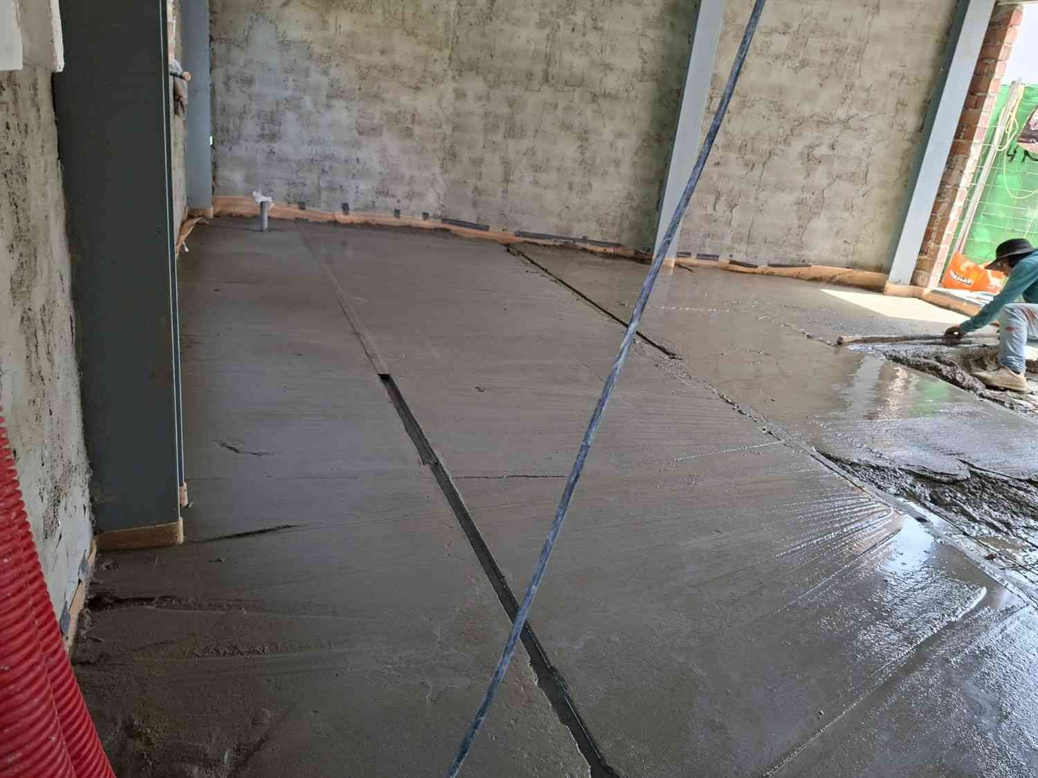 Suelo de hormigón mojado en una habitación sin terminar, con una manguera y obras recientes a la vista.