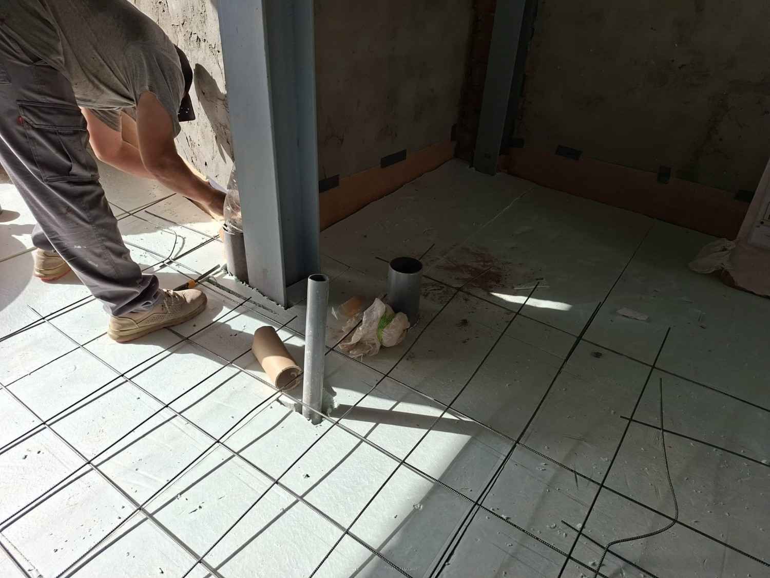 Un trabajador coloca baldosas alrededor de un poste metálico en una habitación en construcción.