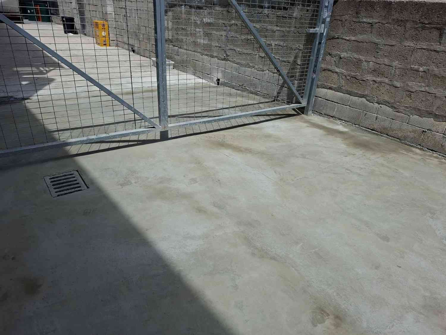 Un patio de hormigón vacío junto a una verja de tela metálica, con sombras proyectadas sobre el suelo.
