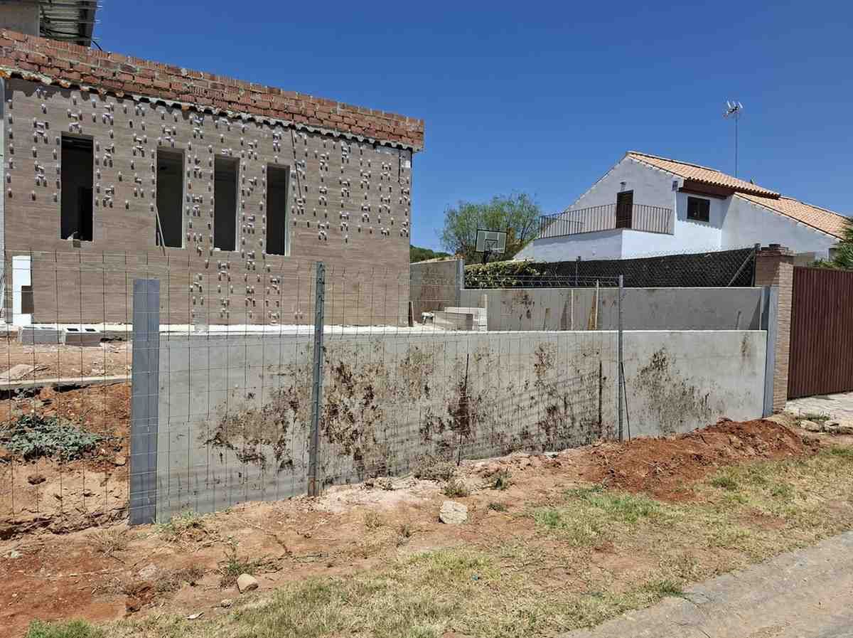 Casa de piedra parcialmente construida y edificio blanco detrás de una valla baja de hormigón bajo un cielo azul despejado.