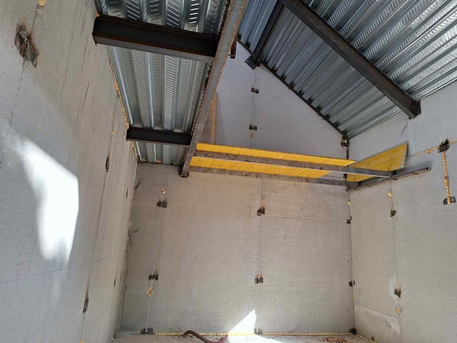 Habitación vacía sin terminar con paredes blancas, techo de chapa ondulada y tuberías o conductos amarillos cerca del techo.
