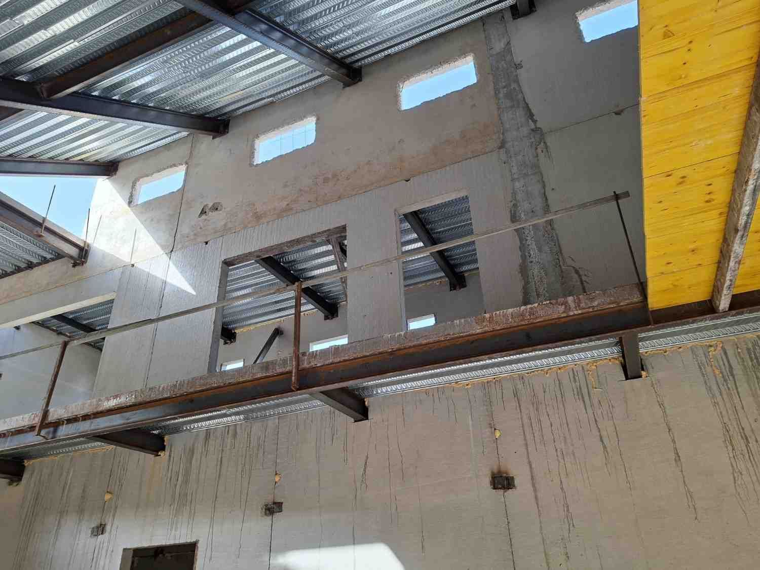 Obra en construcción interior con paredes de hormigón, techo de acero y una viga amarilla en lo alto.