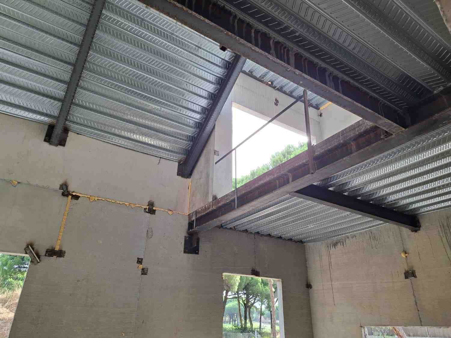 Interior de un edificio en construcción con cerchas metálicas a la vista y paredes de hormigón sin acabado.