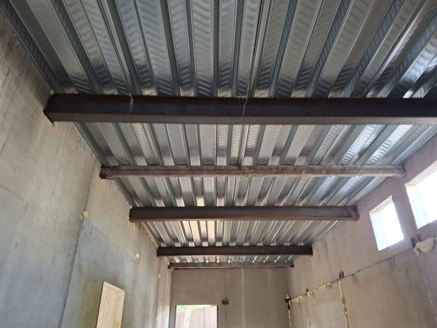 Interior de una habitación con techo de chapa ondulada, vigas a la vista y paredes de hormigón visto.