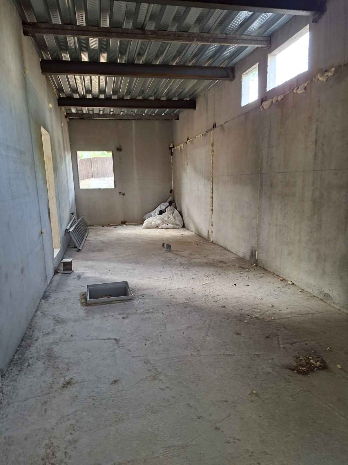Almacén vacío y sin terminar, con paredes de hormigón, techo de metal y escombros en el suelo.