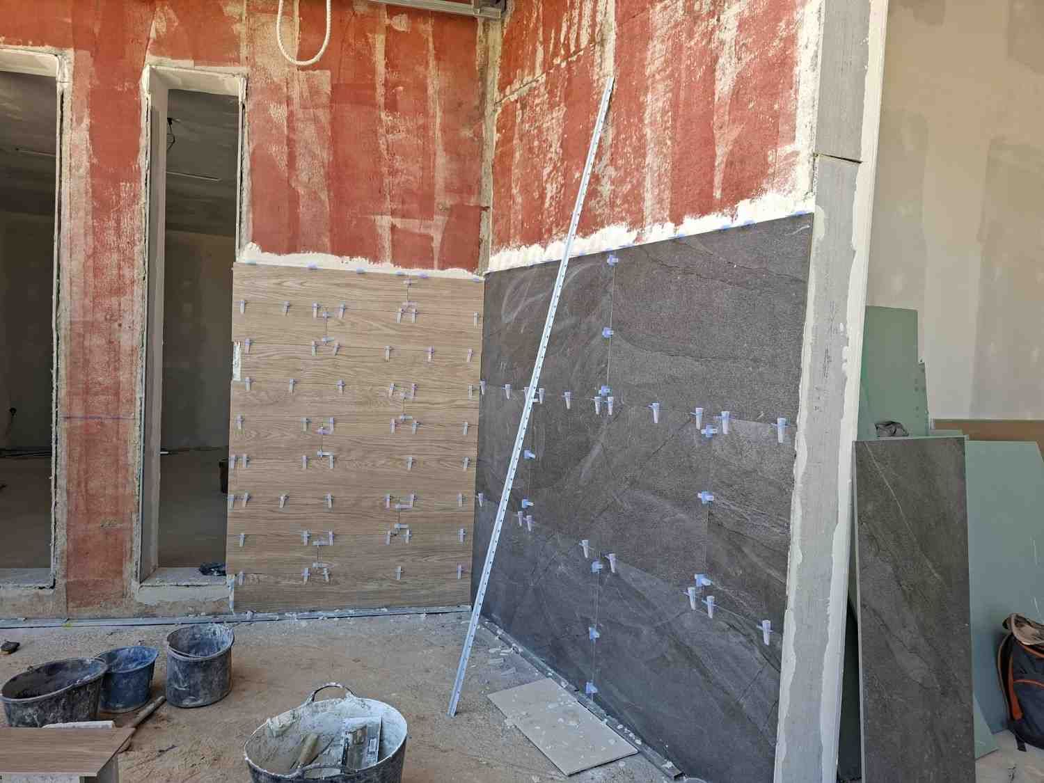Pared interior sin terminar, con yeso y azulejos a la vista; materiales de construcción esparcidos por el suelo.