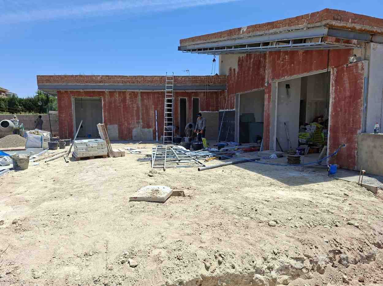 Casa de ladrillo rojo parcialmente construida en un terreno polvoriento, con materiales esparcidos en las cercanías.
