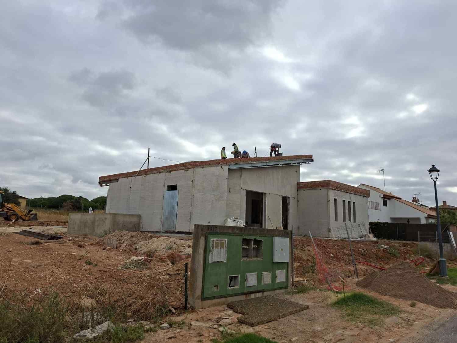 Pequeña casa de hormigón en construcción con trabajadores en el tejado y una caja de servicios públicos verde en la fachada.