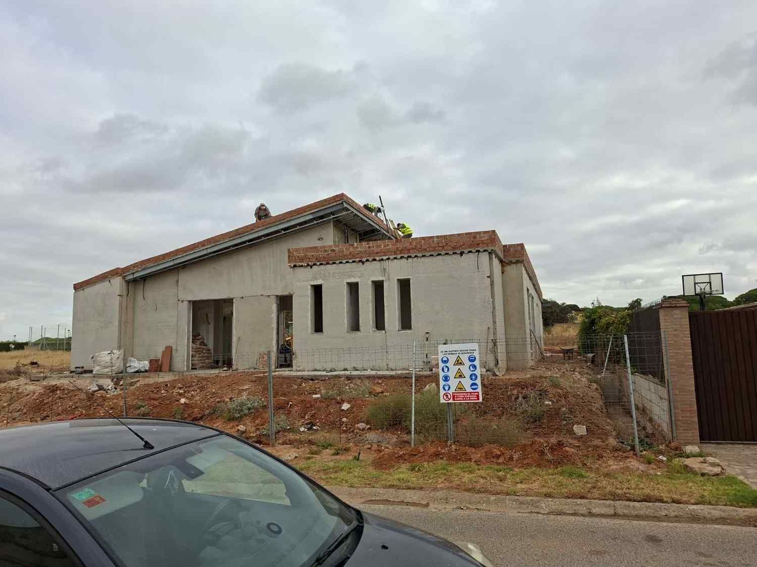 Casa gris parcialmente construida en un terreno de tierra bajo un cielo nublado, con un coche aparcado en primer plano.