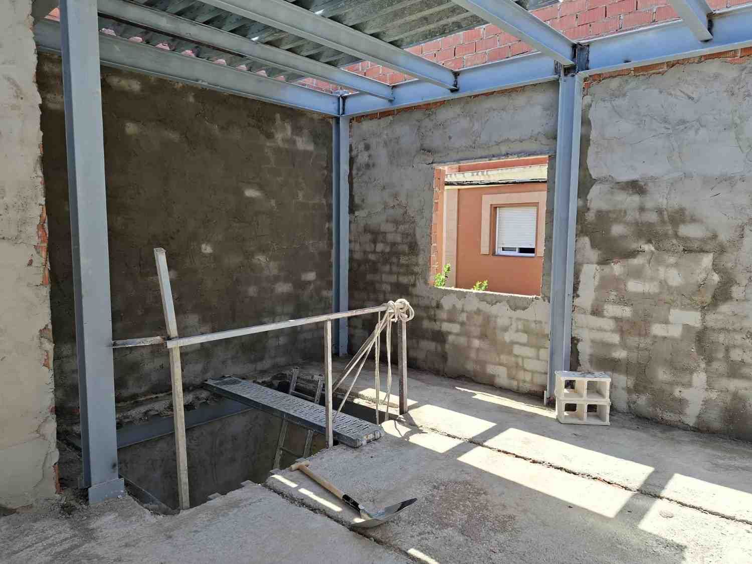 Habitación de hormigón sin terminar con vigas a la vista, escalera abierta y luz solar que entra por una ventana.
