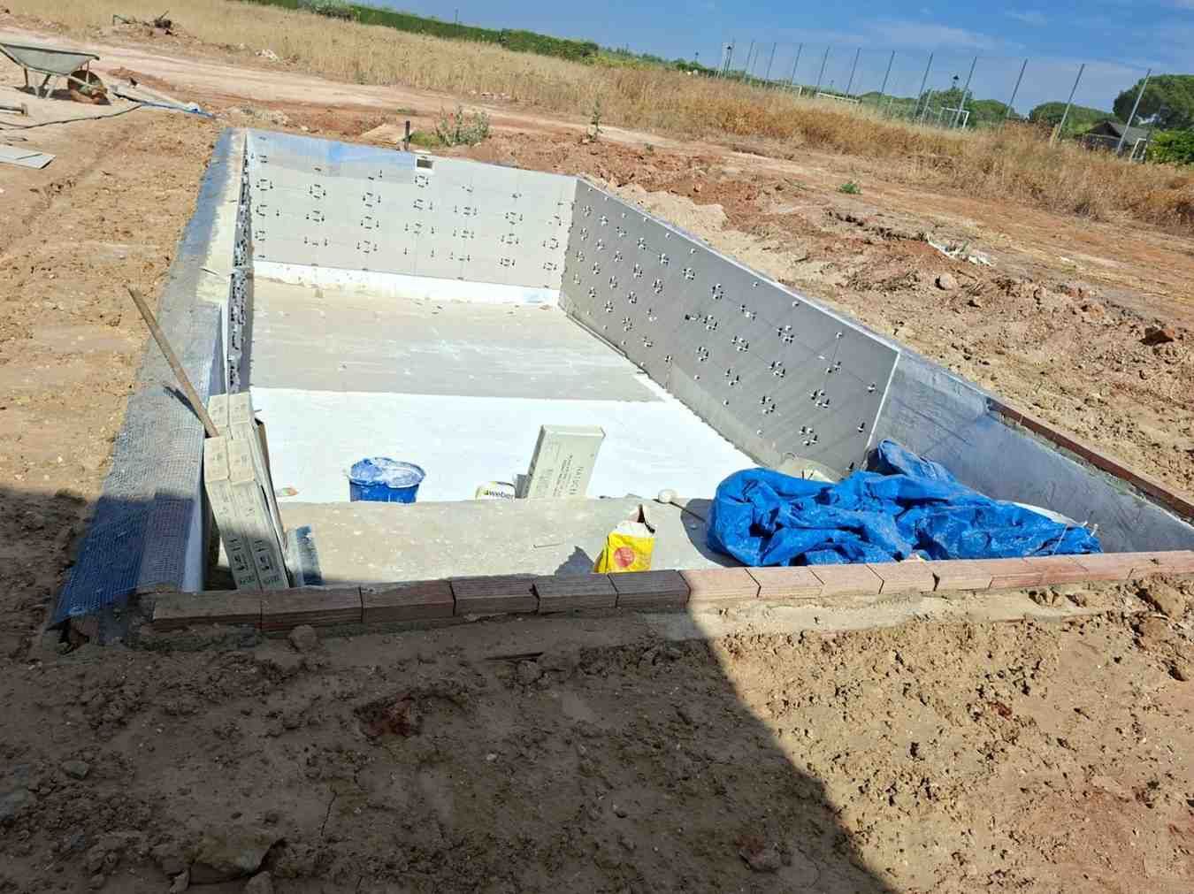 Foso rectangular de hormigón en construcción en un campo de tierra seca, con encofrado metálico y lona azul en el interior.