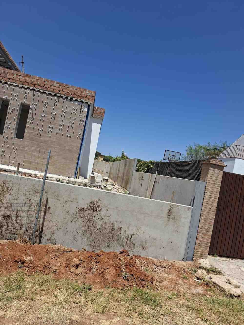 Muro de hormigón parcialmente construido junto a casas de ladrillo bajo un cielo azul despejado.