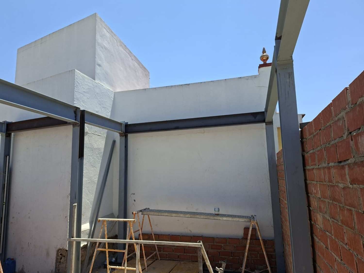 Muro blanco parcialmente construido y estructura metálica en una zona de construcción con andamios y una pared de ladrillo.