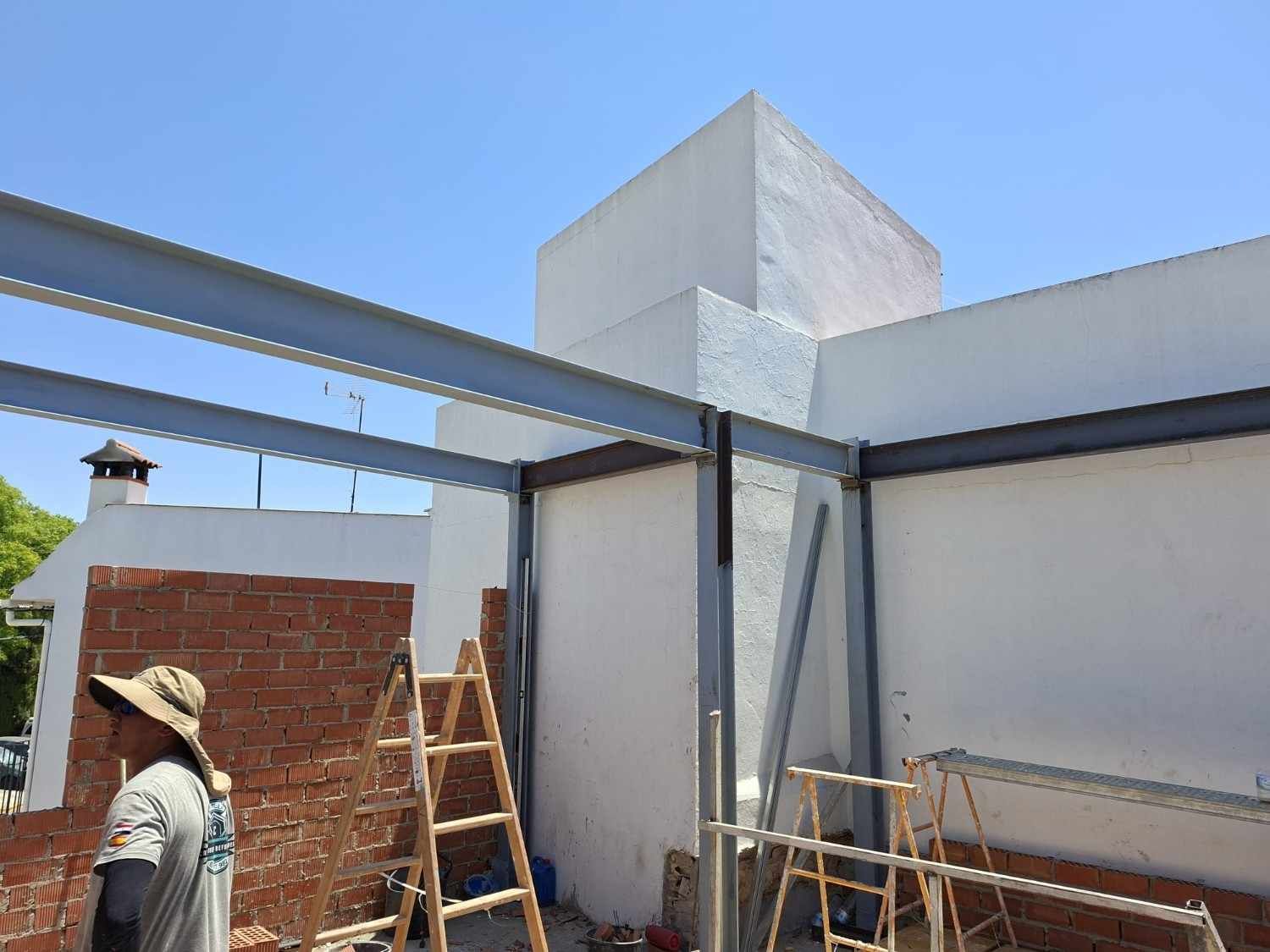 Obrero subido a una escalera junto a un edificio blanco en construcción, con vigas metálicas en el techo.