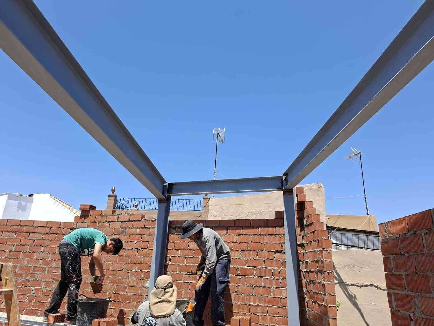Dos trabajadores construyendo muros de ladrillo bajo una estructura de vigas de acero en una obra de construcción.