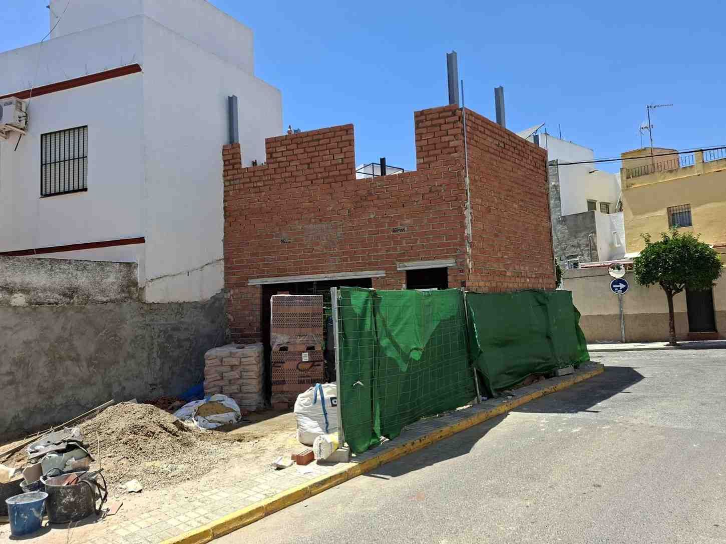 Muro de ladrillo rojo parcialmente construido junto a una casa blanca, cubierto por una valla verde en una calle tranquila.