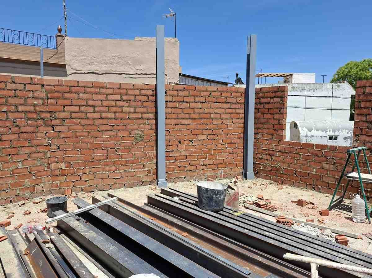 Muro de ladrillo parcialmente construido con postes de hormigón y materiales de construcción bajo un cielo azul.